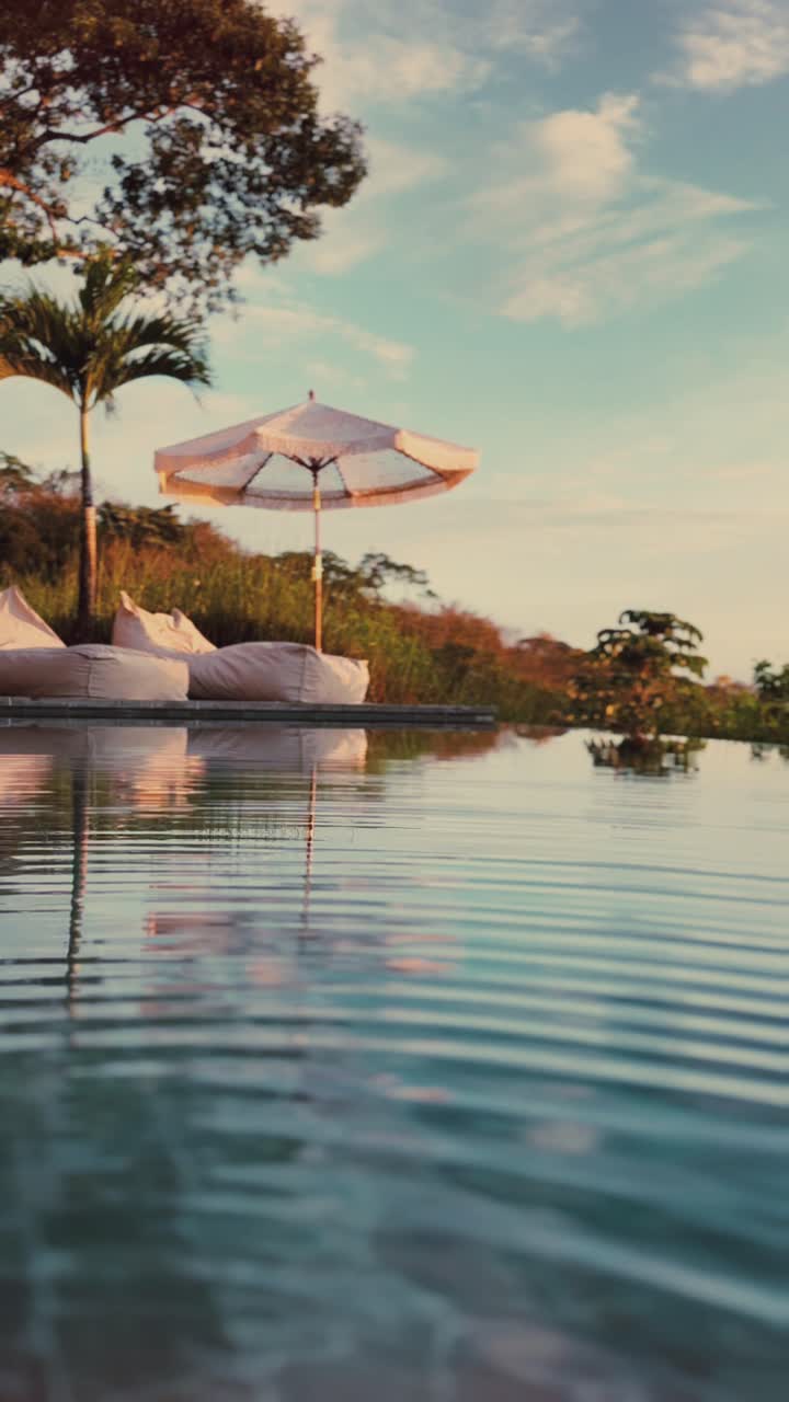 Serene infinity pool reflecting the sky, with a sun umbrella and cozy cushions on the deck, surrounded by lush tropical nature
