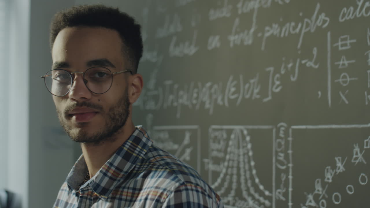A young man in glasses standing in front of a chalkboard filled with scientific equations and graphs
