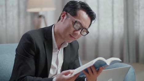 close up of asian businessman in jacket and shorts reading a book after working with a laptop at home