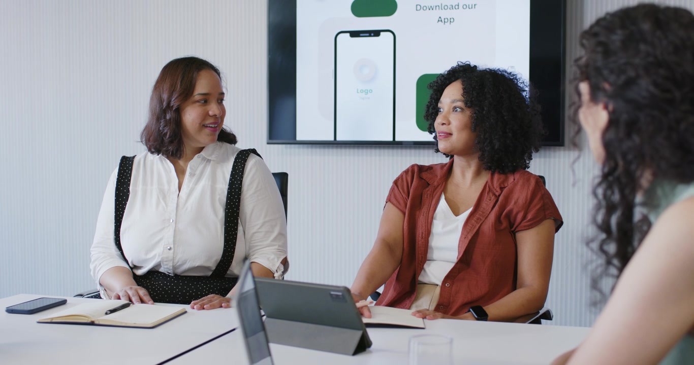 African American women showing app mockup at table, speaker gesturing with tablet, colleague noting