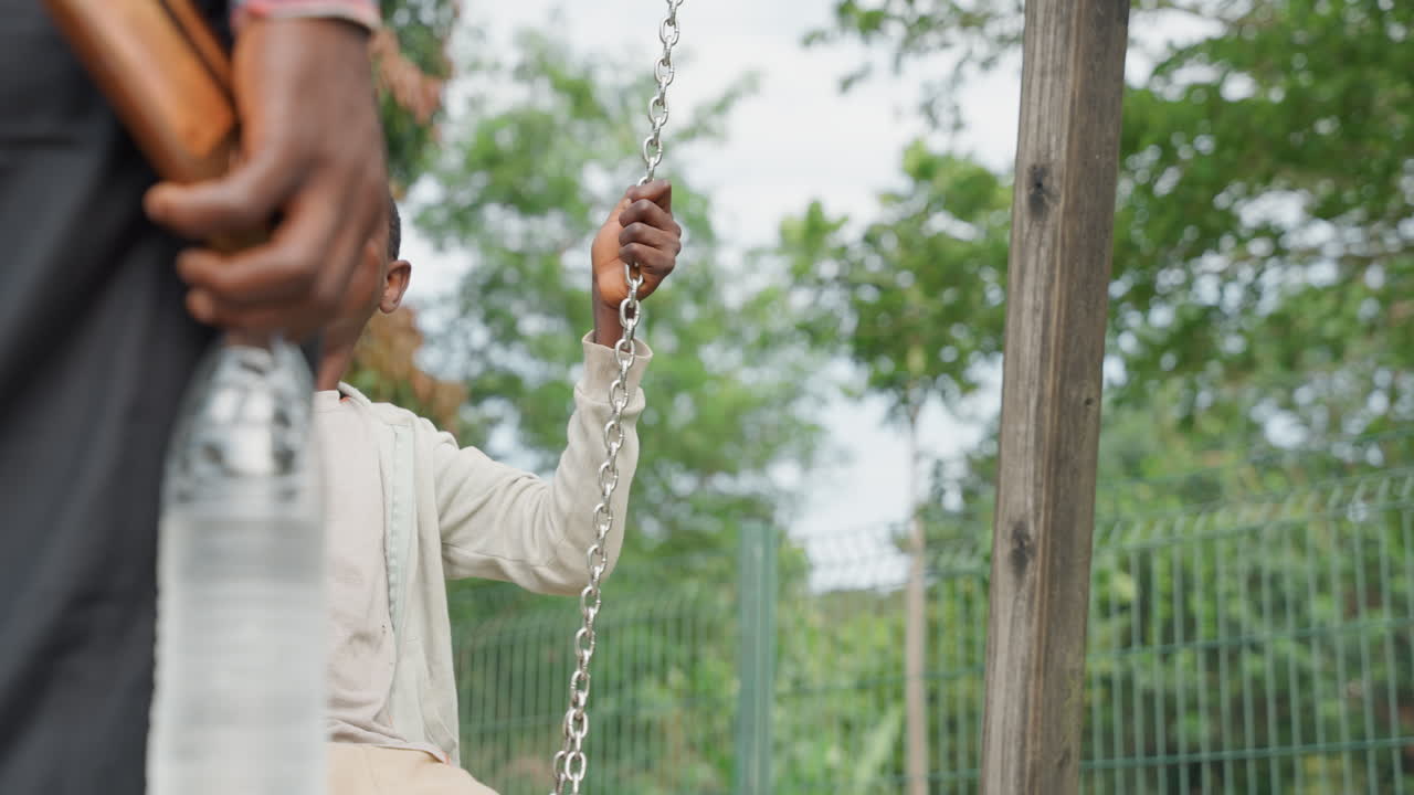 Black father pushing son on swing, laughing expression, tall trees, bottle in hand, closeup low angle, motion blur, active parenting, warm daylight, playground fence, carefree energy, weekend bonding