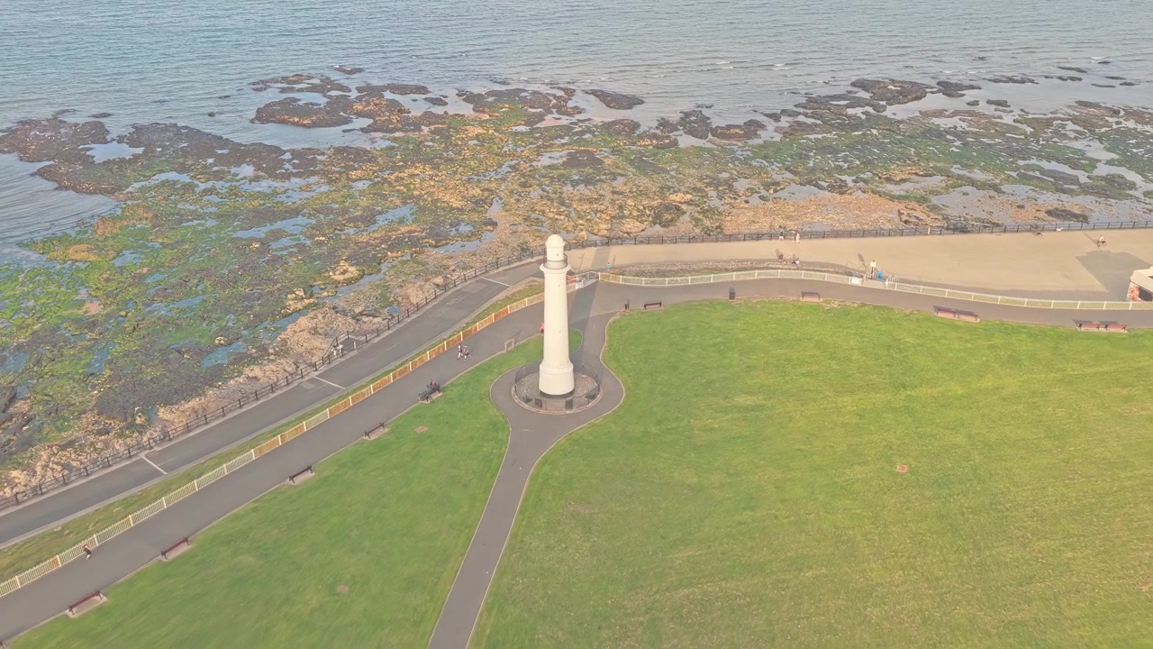 Il faro di Roker si erge accanto al Mare del Nord lungo la Roker Promenade a Sunderland, Tyne and Wear, affacciandosi su una costa rocciosa intertidale e un parco costiero, discesa del drone con movimento in avanti.