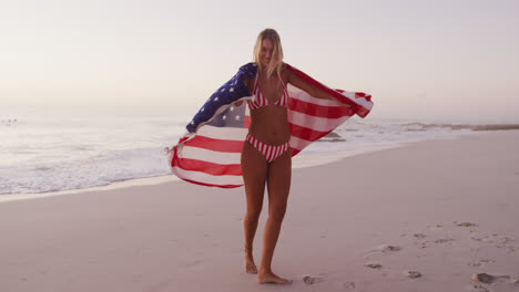 una mujer caucásica sosteniendo y agitando una bandera estadounidense en la playa.