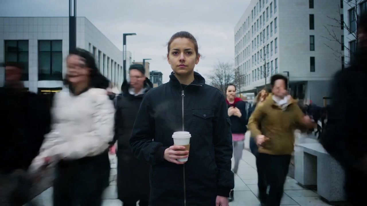 Standing woman in dark jacket holding steaming to-go cup on office plaza, commuters rushing past