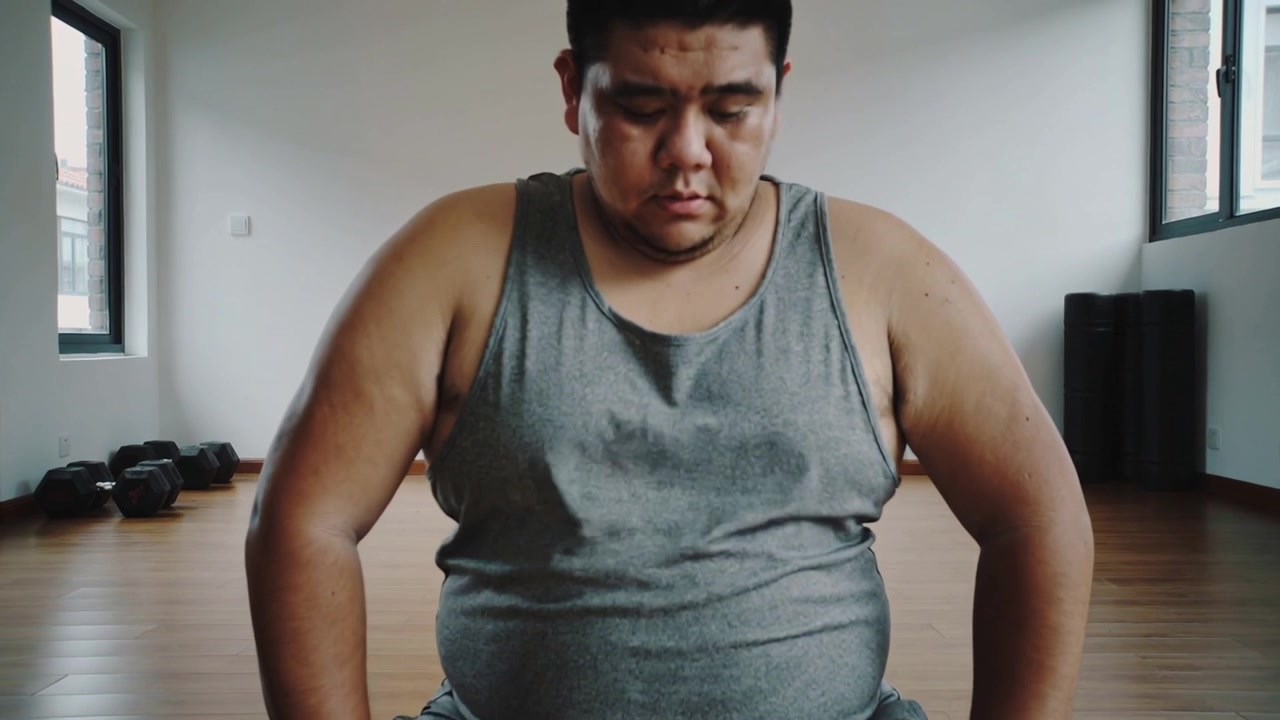 Hombre sentado ajustando la postura en la colchoneta de ejercicio después de una serie, vistiendo una camiseta sin mangas gris, con mancuernas cerca.