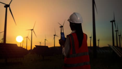 back view of asian female engineer in a helmet looking at the wind turbine blueprint on smartphone while standing in front of wind turbines rotating at sunset