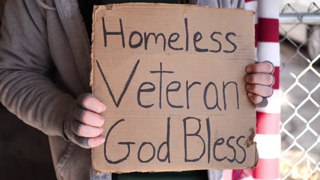 Homeless veteran man holding sign close up that says "Homeless veteran, God bless" with rolled up American flag behind him