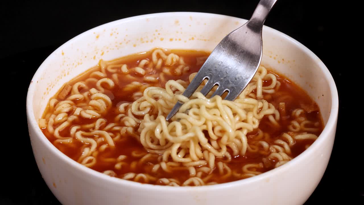 A hand uses a fork to twirl and lift cooked instant noodles from a steaming bowl of soup, shot in close-up with bright, even lighting