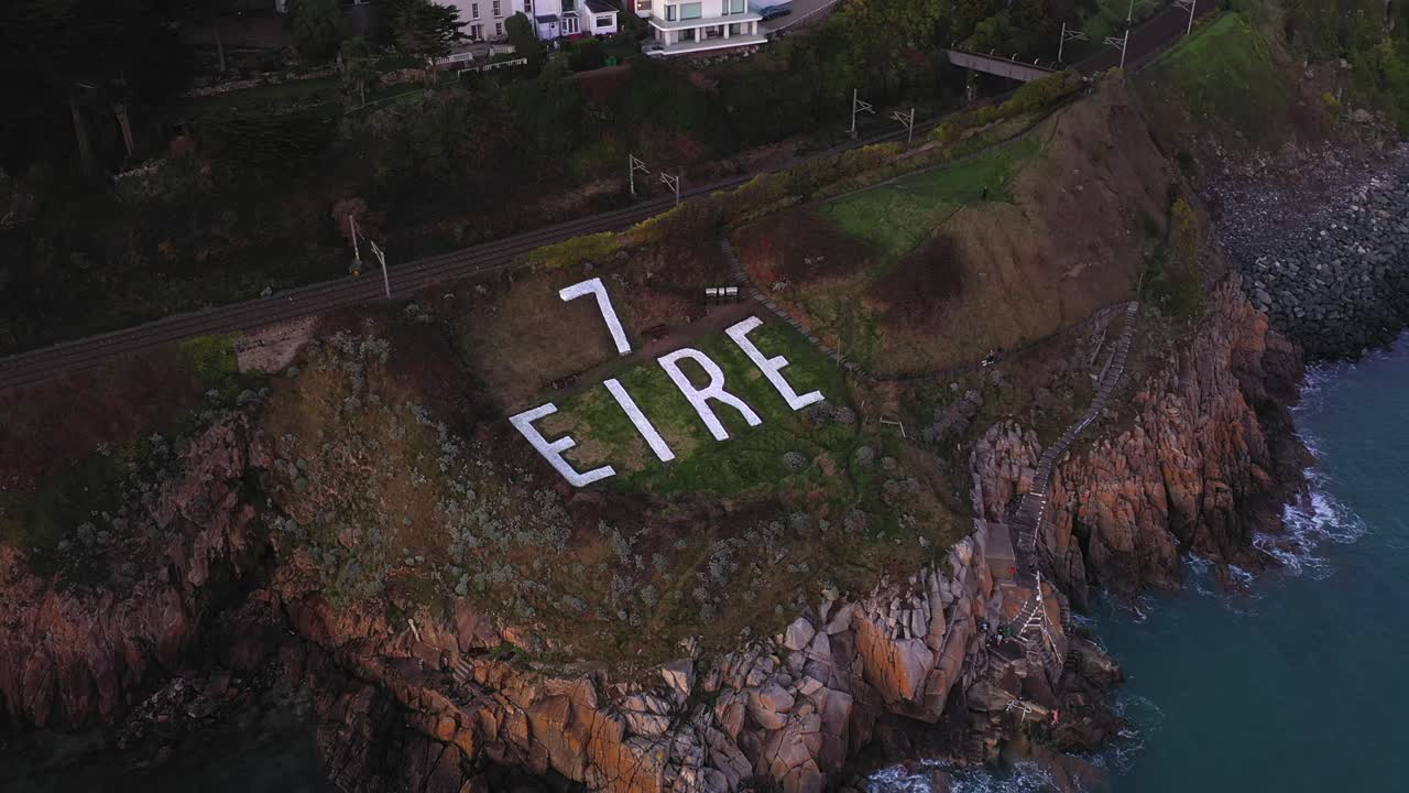 Eire 7, World War II Coastal Watch Marker Sign, Killiney, Dublin, Ireland. January 2020. Drone descends gradually revealing people exercising, walking dogs and swimmers at base of the coastal cliffs
