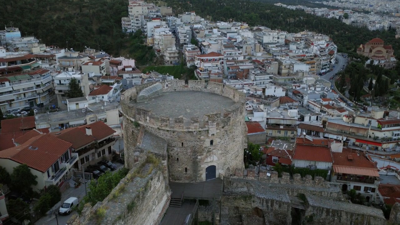 Vista aerea con drone all'alba delle torri della fortezza di Eptapirgio e delle antiche mura di Salonicco, che si affaccia su case dai tetti rossi e sulla Chiesa di San Paolo.