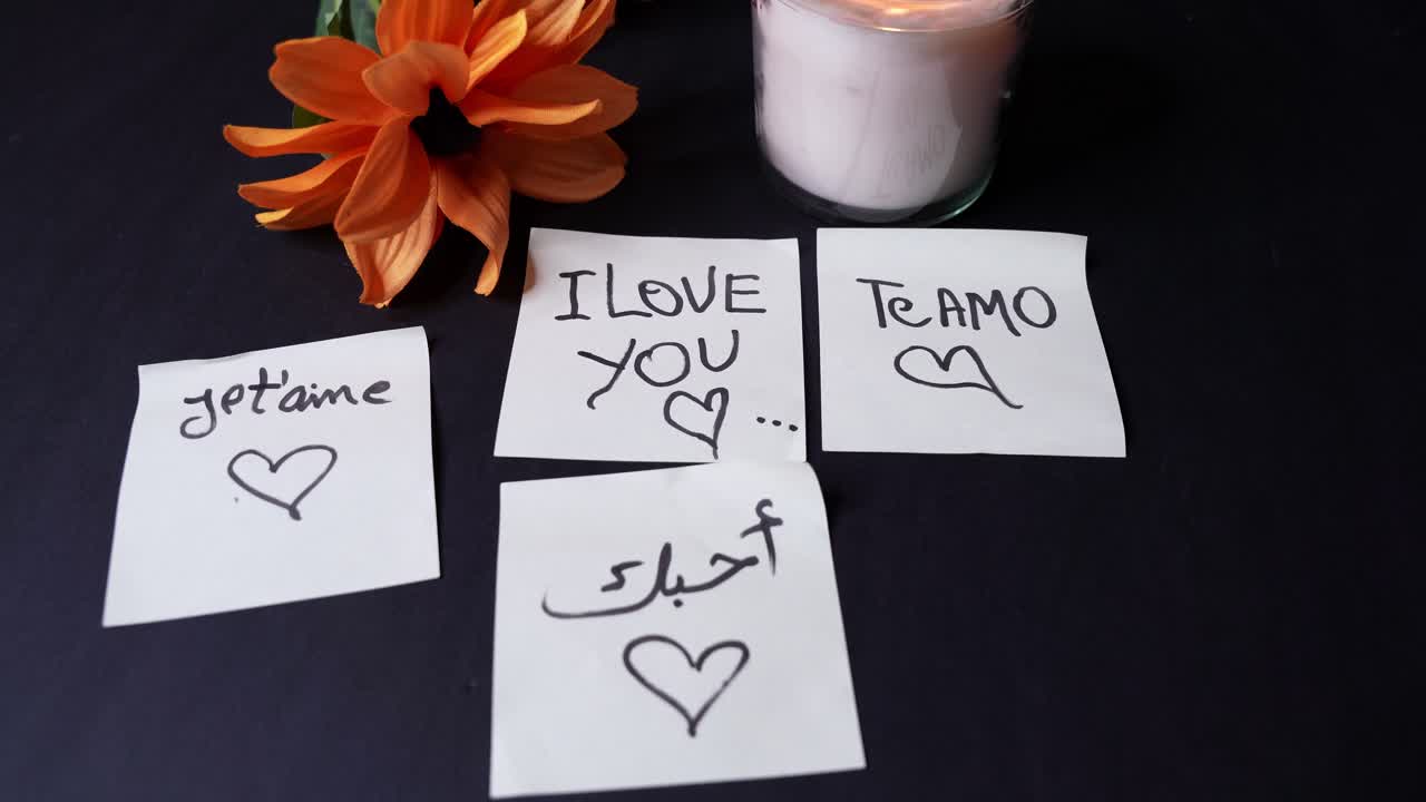 Overhead nærbillede af et tændt glaslys, orange blomster og sticky notes, hvor der står “Jeg elsker dig” på flere sprog på en mørk bordplade, hvilket skaber en varm, romantisk indendørsstemning