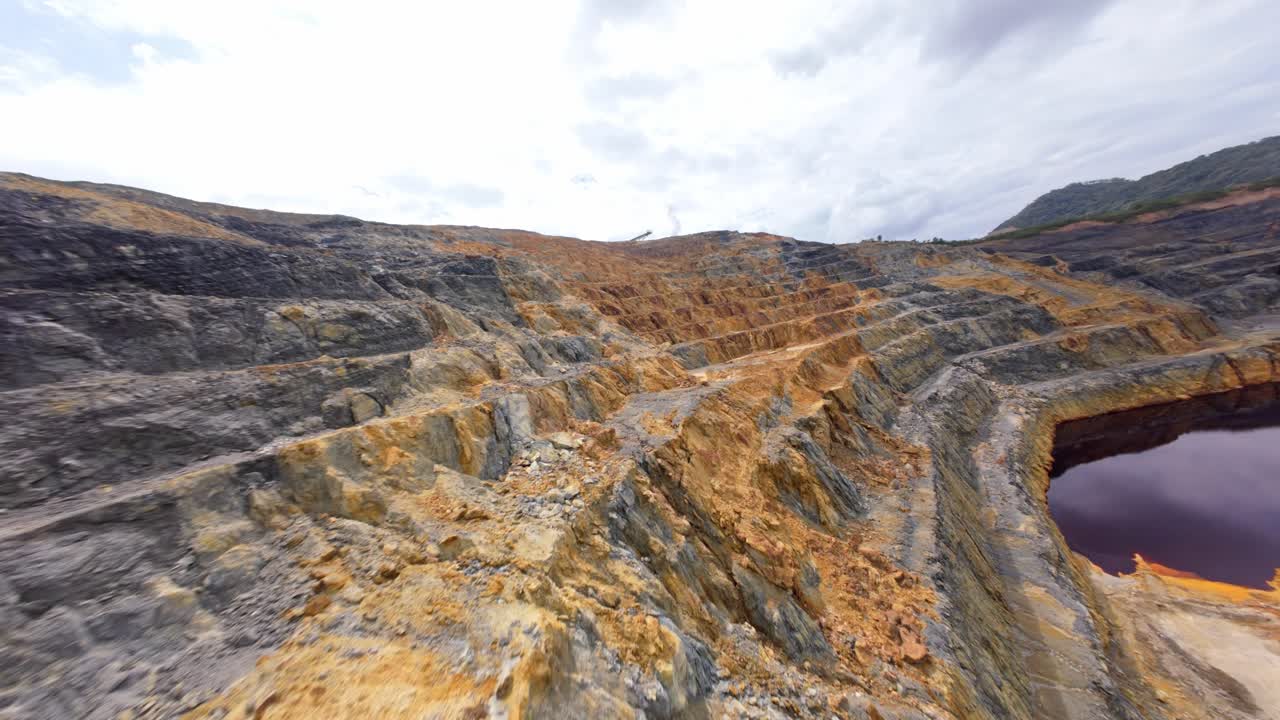 Colorful terraced rock walls of an open-pit mine excavation, dark tailings pond, Barrick Gold, Cotuí, Mining industry, environment, Dominican Republic. Aerial drone FPV, low flight