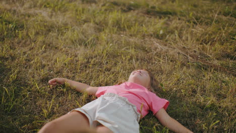 young girl in pink shirt lies on grass with arms spread, gazing up at sky in peaceful countryside, soft golden sunlight highlights her relaxed posture, with cousin partially visible in foreground