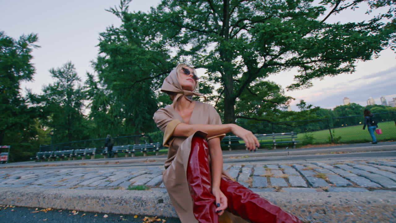 New York, AS, 5 Oktober 2025: Wanita berkerudung sutra berpose di Central Park. Profil samping wanita mengenakan kacamata hitam dan kerudung krem duduk di luar ruangan di taman kota.