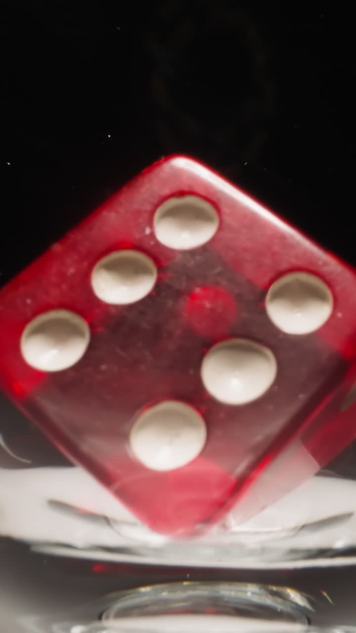 Red gaming cube with white dots floating in transparent wineglass at studio light on black background. Dice for gambling casino games with six points on each face macro