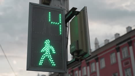 close view of a digital traffic light showing a countdown with a green pedestrian signal, with a red city building and a cloudy sky