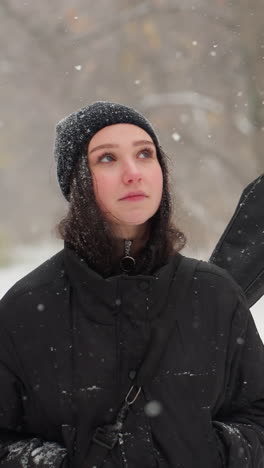 dama con capucha negra llevando una guitarra con las manos en los bolsillos caminando pensativamente bajo copos de nieve, banco nevado en el fondo añade a la atmósfera serena del invierno