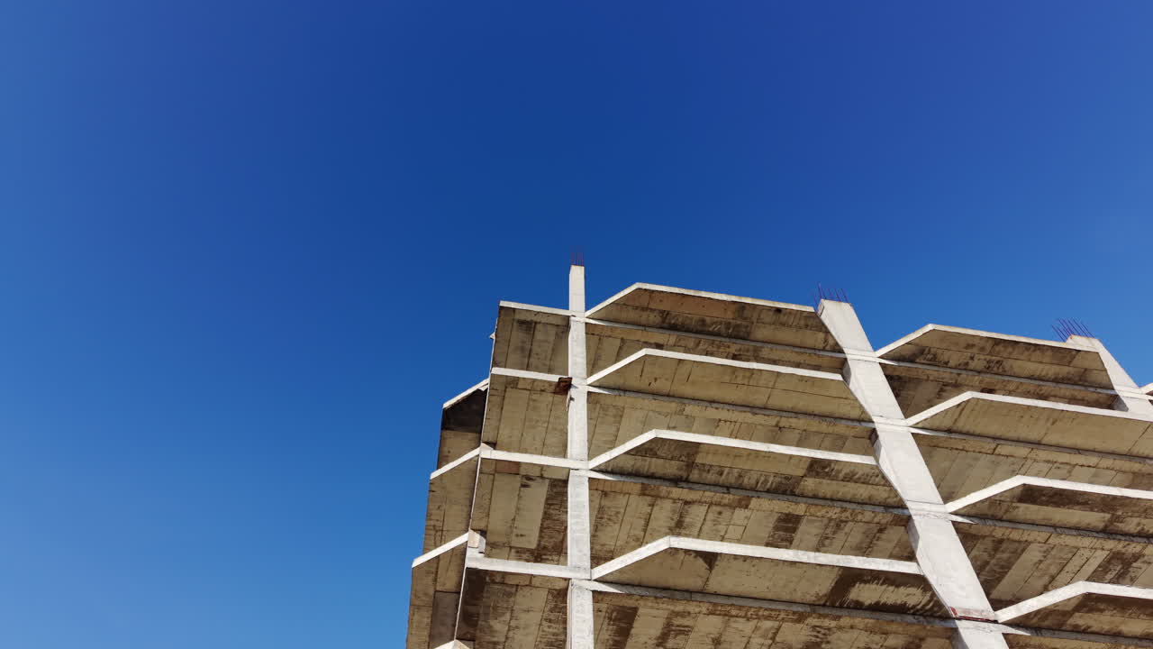A low-angle side view showing multiple levels of a reinforced concrete building frame under construction against a clear blue sky. Aerial