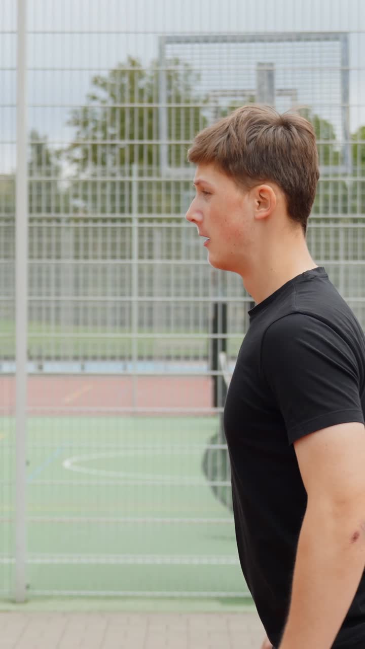 Young man performing bodyweight squat exercise on outdoor sports court, focusing on strength, fitness and training, highlighting healthy and active lifestyle, vertical follow camera shot, slow motion