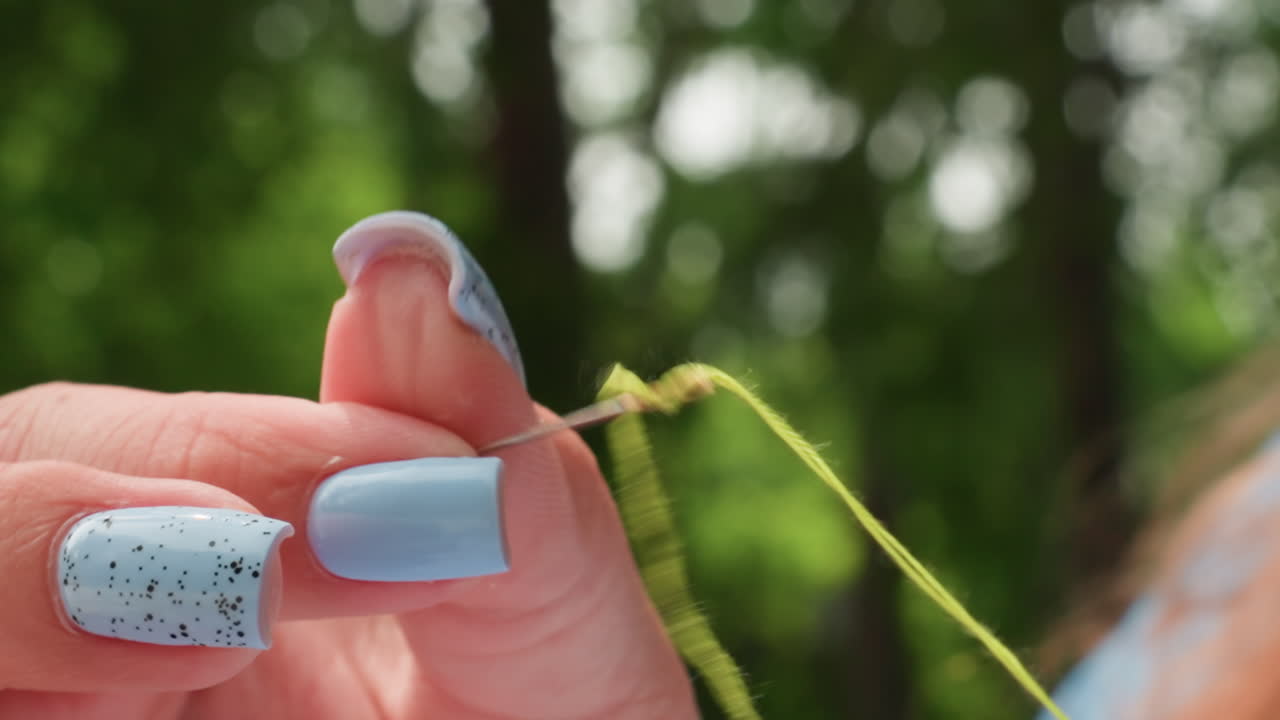 Close up female hands with blue nails threading needle outdoors, green strand guided toward eye, steady fingers and patience, craft preparation in park under soft daylight