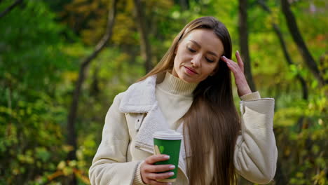 attractive brunette lady in white warm jacket has a walk in the park. woman enjoying coffee and warm sunny autumn weather.