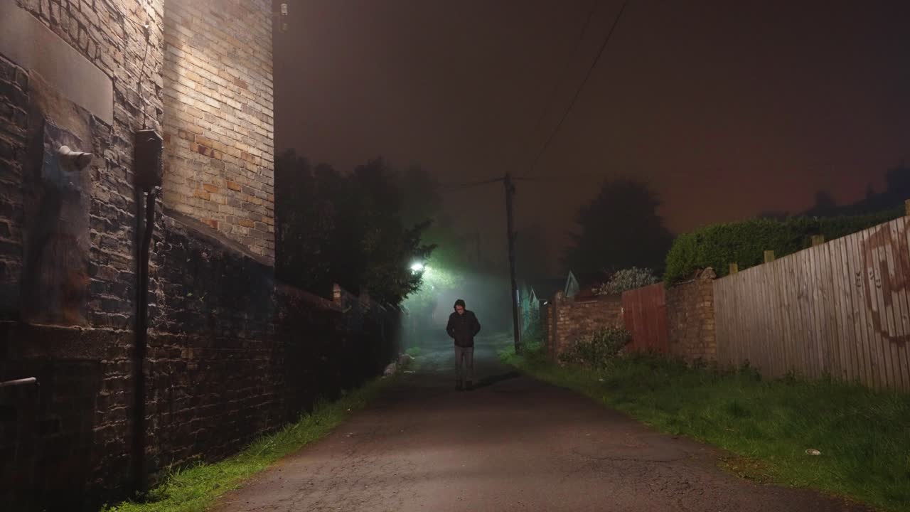 Hooded man in dark jacket walking down foggy alley at night, locked off