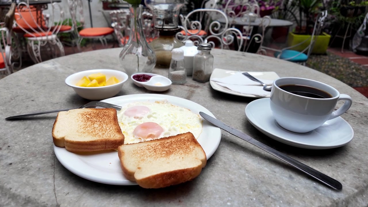 Tampilan dekat hidangan sarapan khas yang menampilkan telur goreng dan roti panggang disajikan dengan kopi Kolombia segar di teras luar ruangan.