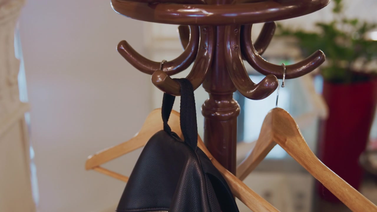 Primo piano di una persona che appende uno zaino in pelle nero su appendini in legno attaccati a un appendiabiti marrone in un ambiente interno. Una mano con manica scura si allunga verso il gancio e posiziona la borsa in modo sicuro tra le altre
