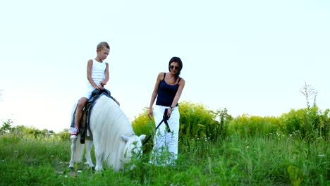 a woman and a boy are walking around the field, son is riding a pony, mother is holding a pony for a bridle. cheerful, happy family vacation. outdoors, in summer, near the forest