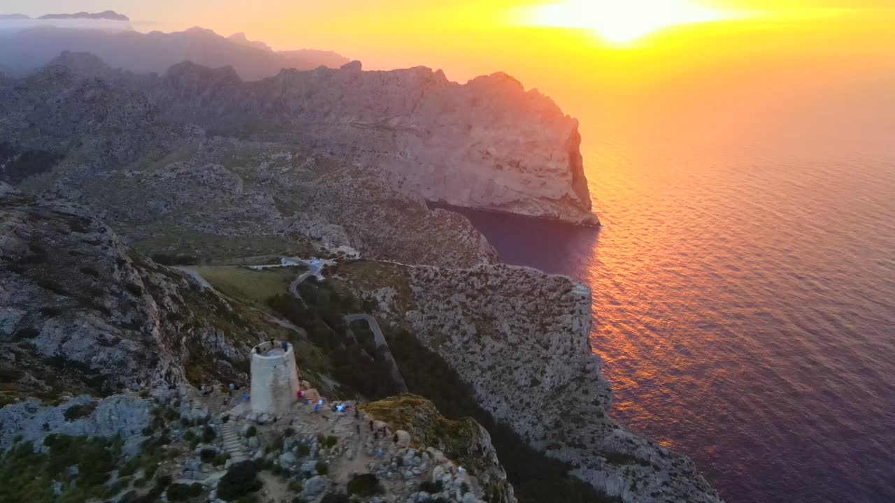 Experience the stunning sunrise over the iconic Mirador es Colomer in Pollensa, Mallorca. The serene landscape features rocky cliffs, sparkling waters, and a historic tower.