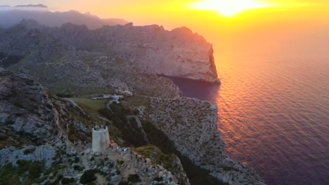 experience the stunning sunrise over the iconic mirador es colomer in pollensa, mallorca. the serene landscape features rocky cliffs, sparkling waters, and a historic tower.