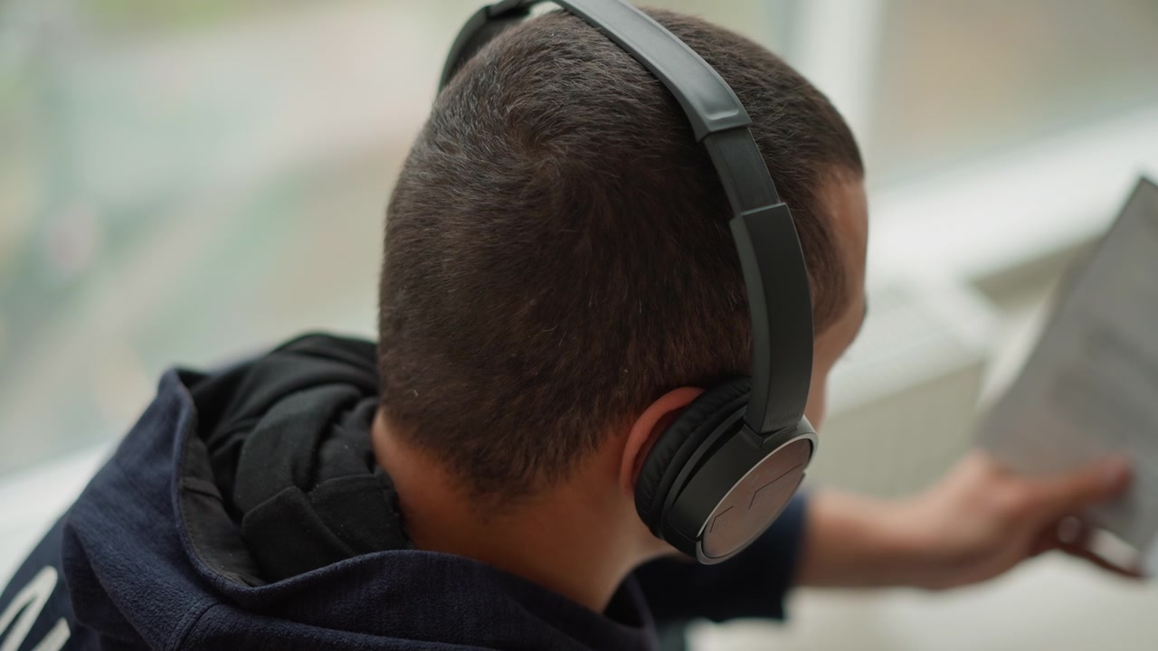 Primo piano di un giovane ragazzo che indossa cuffie nere e una felpa con cappuccio blu scuro, che tiene in mano un foglio mentre guarda in basso. Il ragazzo rimane fermo con un leggero movimento della mano mentre interagisce con il documento.