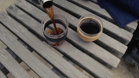 Woman-Pouring-Hot-Coffee-From-Thermos-Into-glass-Mug-On-Wooden-bank-at-the-beach-in-slow-motion