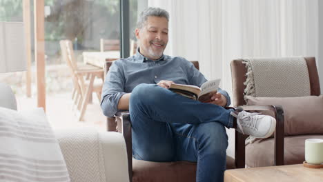 Glücklicher-älterer-Mann-Mit-Gemischter-Abstammung,-Der-Im-Sessel-Sitzt-Und-Im-Wohnzimmer-Ein-Buch-Liest,-Zeitlupe