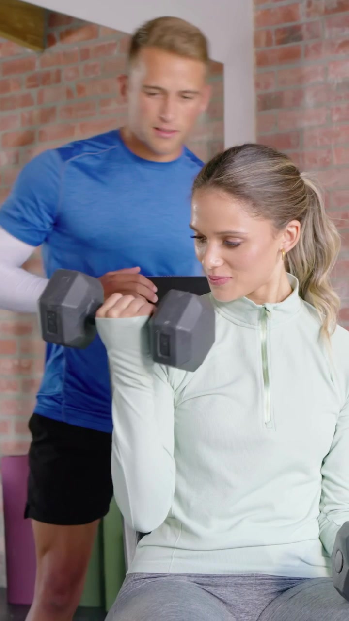 Vertikal video: Træner i blå skjorte griber hex-håndvægt i fitnesscenter, spotter klient og korrigerer form.