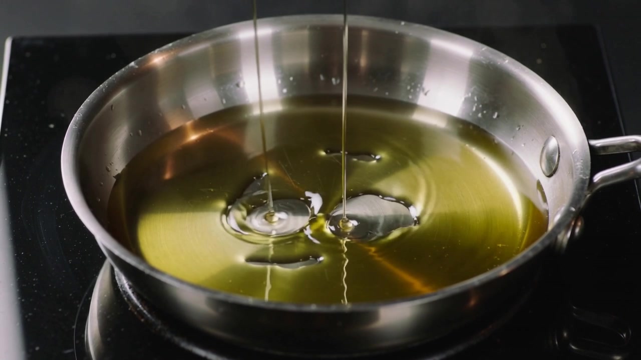 Olio che cola dall'alto formando pozze in una padella di acciaio inossidabile su un piano cottura lucido, ricoprendo la padella.