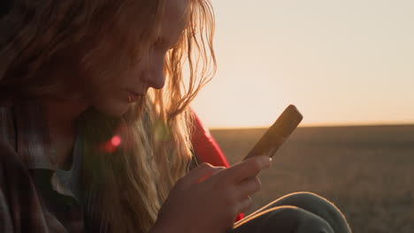 A-teenage-girl-sits-in-the-trunk-of-a-car,-uses-a-smartphone.-Against-the-backdrop-of-a-rural-landscape-where-the-sun-sets