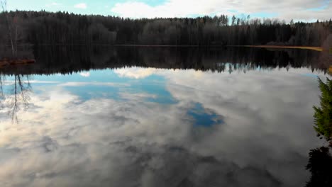 flying at low altitude over a small lake with green trees, a yellow field and the blue sky with the clouds reflecting in the water