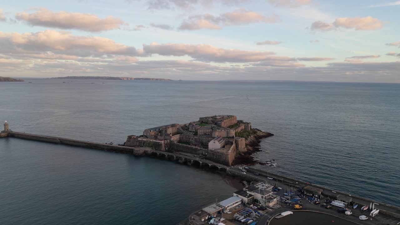 Ampia vista di Castle Cornet, frangiflutti, ingresso del porto e laghetto per modellini di barche a St Peter Port, Guernsey, all'ora d'oro con mare calmo, nuvole rosate e viste verso Jethou