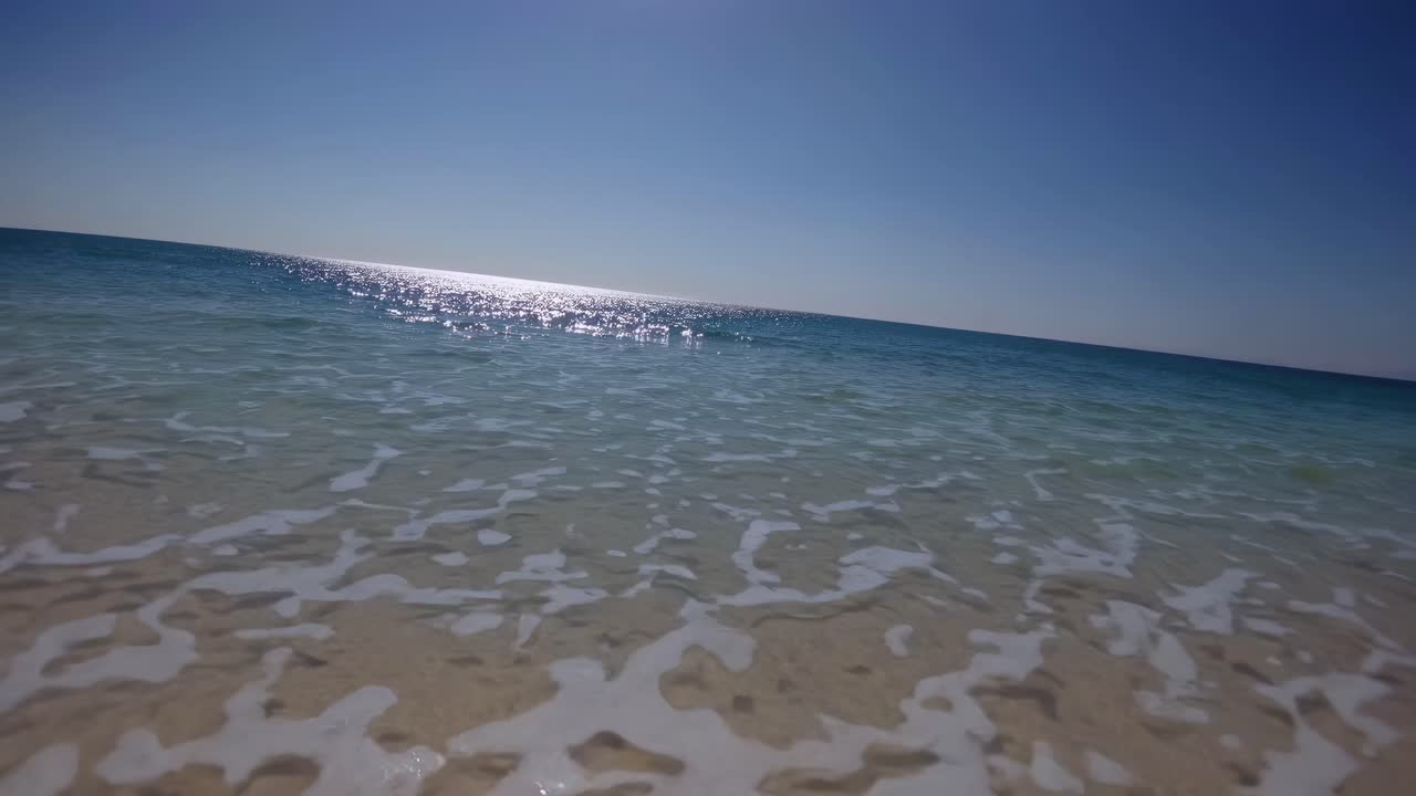 Low-angle video capturing a serene beach scene with gentle waves lapping at the shore under a clear
