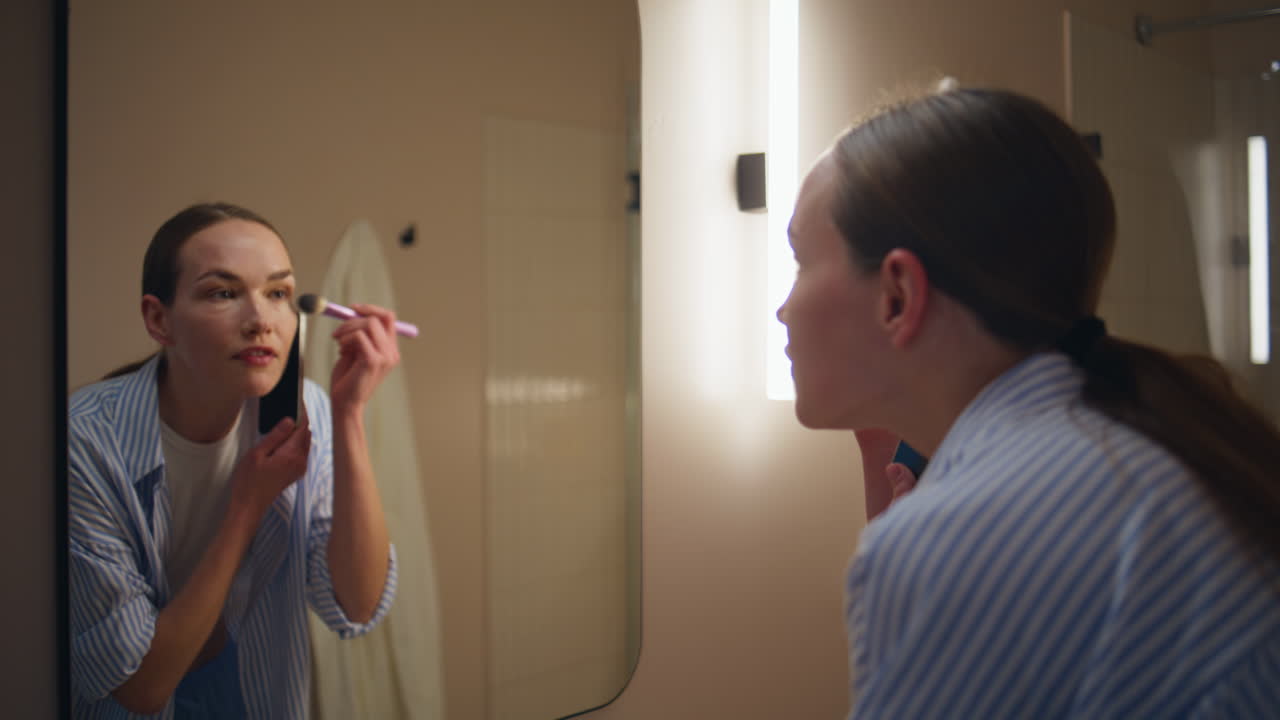 Late girl applying blush talking mobile phone in bathroom closeup. Pretty lady