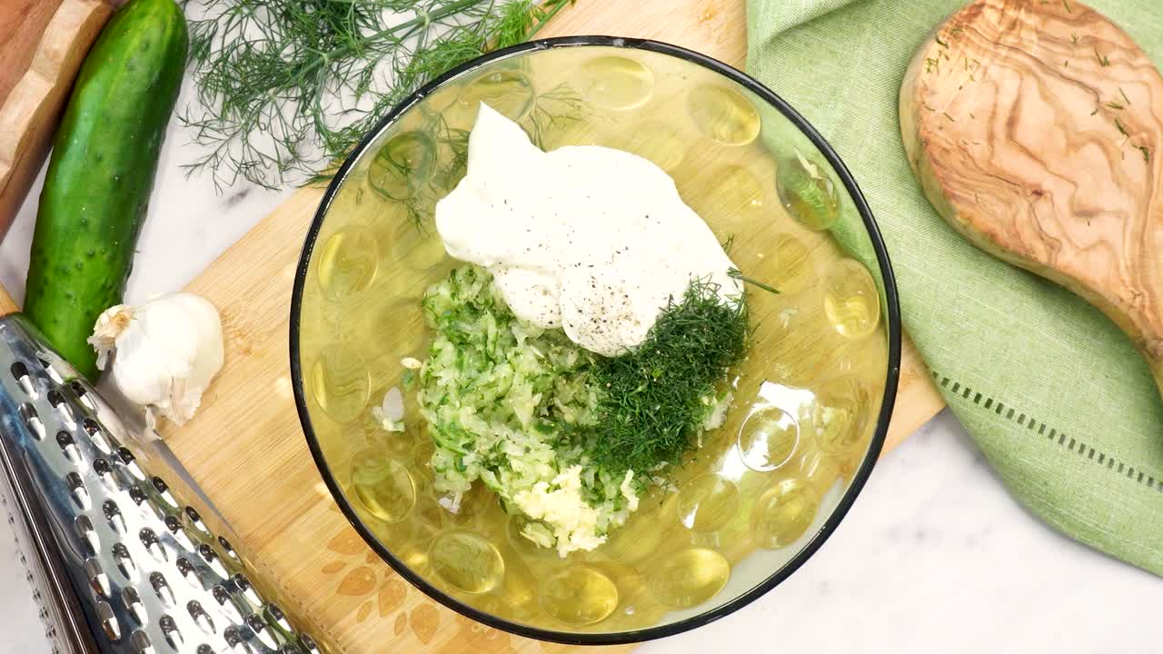 Making Tzatziki Greek or Mediterranean sauce or dip for a party. Dip or sauce in a green bowl on a wooden cutting board with dill, green napkin and a grater. Squeezing lemon juice into the Tzatziki.