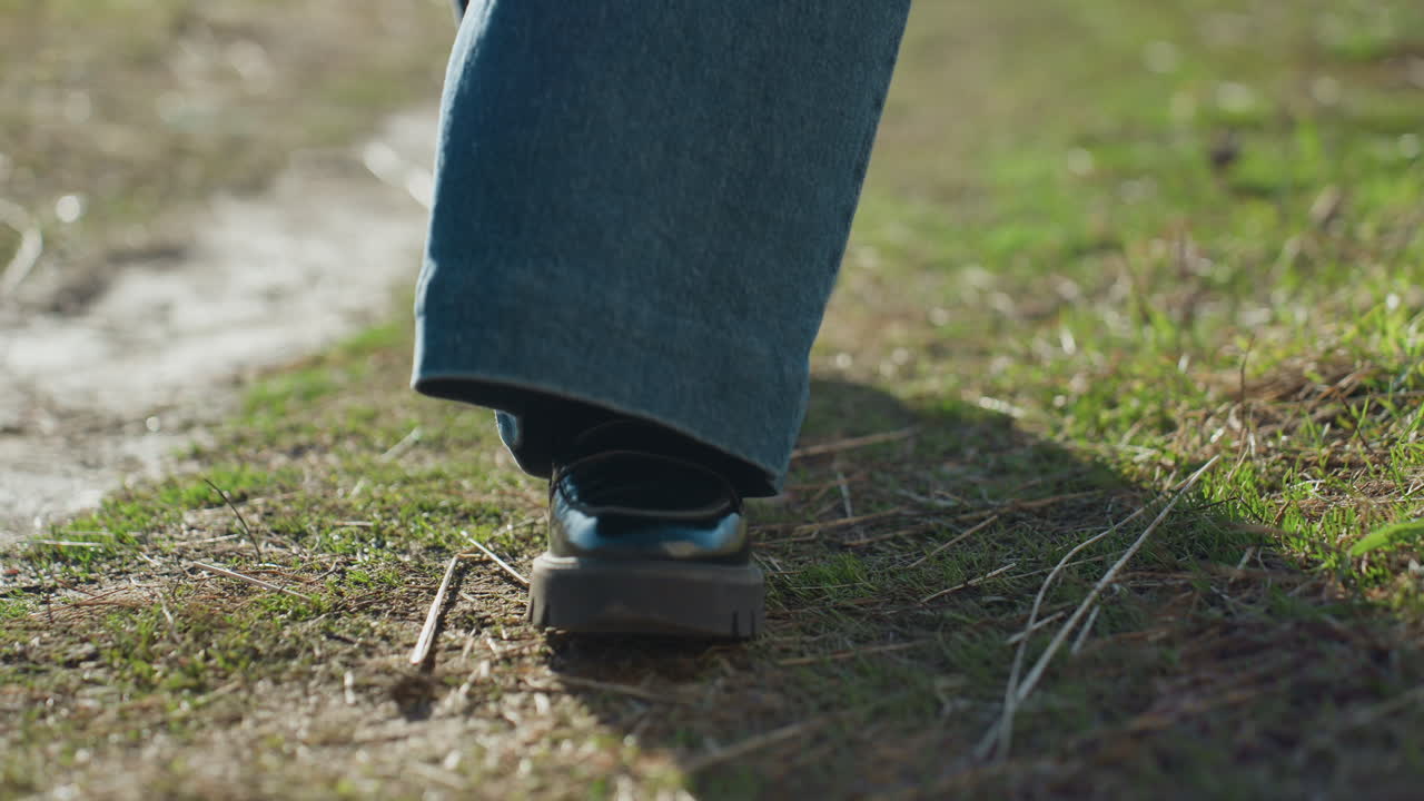 Close-up of person walking outdoors on grassy path with denim jeans and black shoes, sunlight casting shadow on ground, detail of steps emphasizing casual movement and natural environment