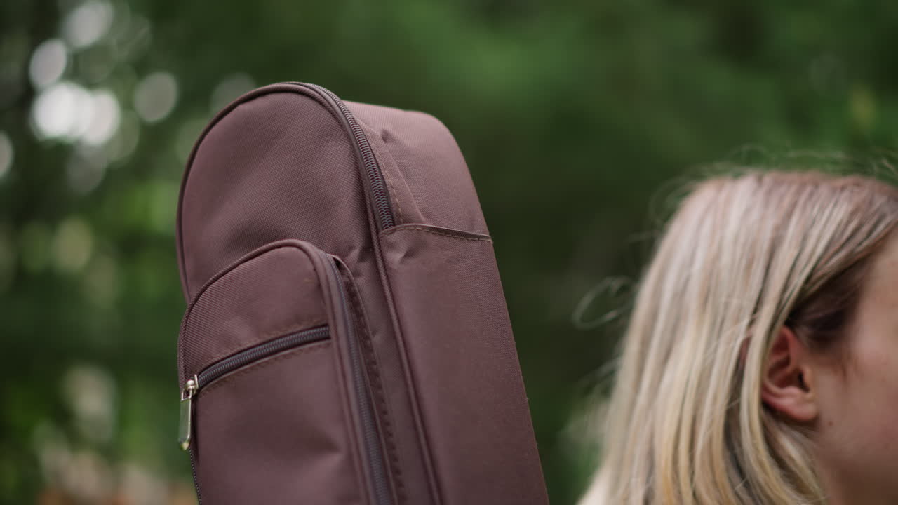 guitar case in park closeup, zipped leather case held over shoulder, blonde musician walking through trees, textured fabric and metal zipper details, blurred bokeh background, movement