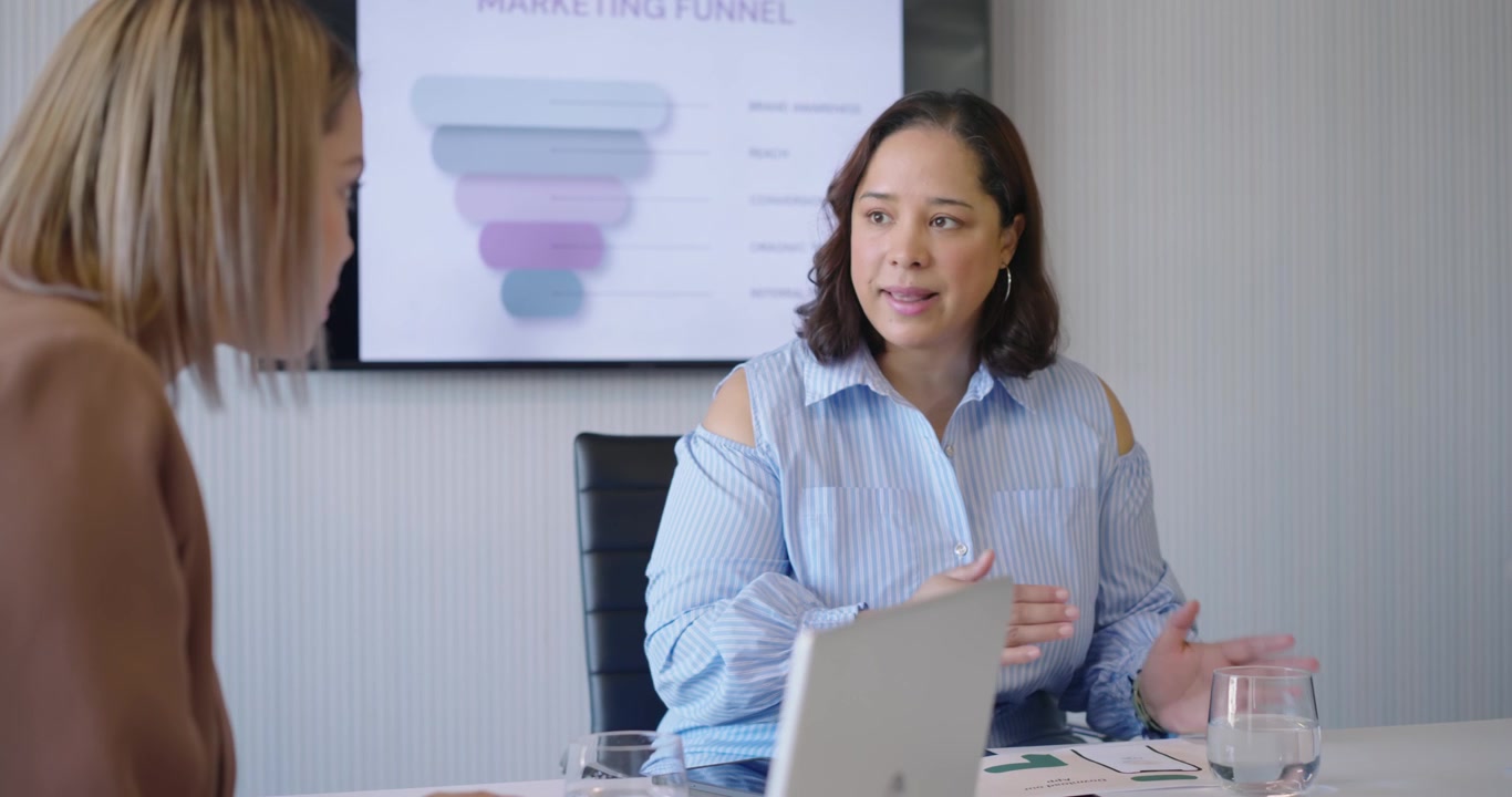 Women coworkers arranging reports, presenting funnel at table, pointing to laptop