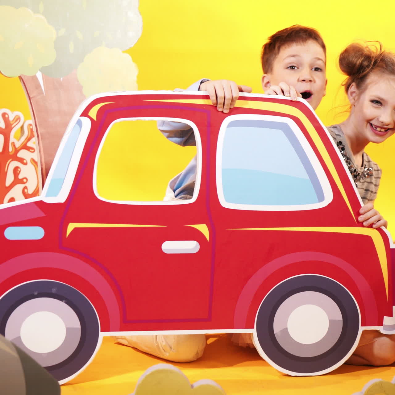 Happy boy and girl playing together in the children's studio room with decor over the yellow background. Beautiful toyful carton scenery of a big red car and colorful bushes.