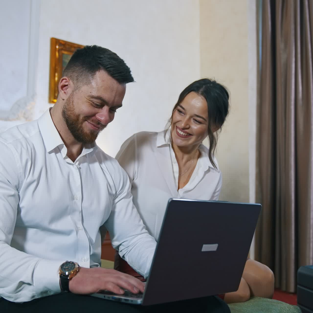 Young couple on trip wedding. Happy guy and girl sitting together on a bed and looking into the laptop in a hotel room. Man and woman at business trip