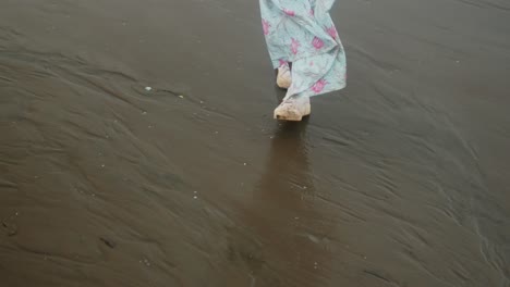 a woman in a light blue dress and sneakers walks on a wet beach in a close-up shot