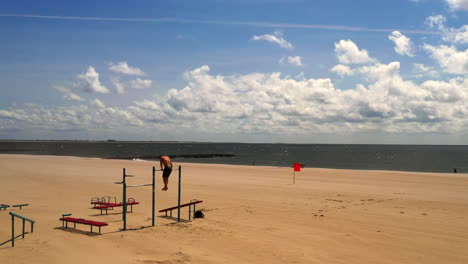 Drohnenkamera-Mit-Stabiler-Aufnahme,-Während-Ein-Junger-Mann-An-Einer-Klimmzugstange-Am-Strand-Von-Coney-Island-In-Brooklyn,-New-York,-An-Einer-Trainingsstation-Trainiert,-Mit-Dem-Sandstrand,-Dem-Atlantischen-Ozean,-Dem-Blauen-Himmel-–-Horizont-Im-Blick