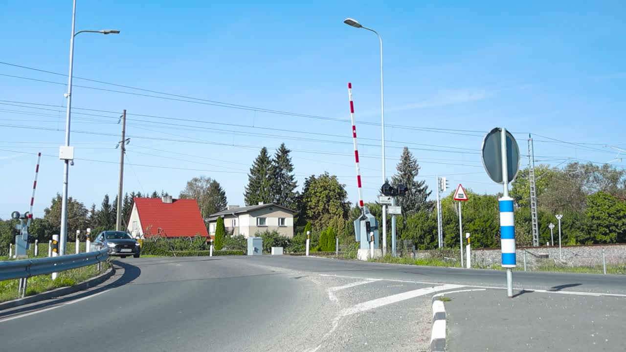 Railway or a railroad crossing with asphalt road that has marking on it and boom gates up during summer time while sun is shining. Cars driving in slow motion and blue sky and trees in the background.